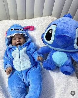 Smiling baby in a blue Stitch-style onesie lying next to a large blue plush toy on a white blanket.