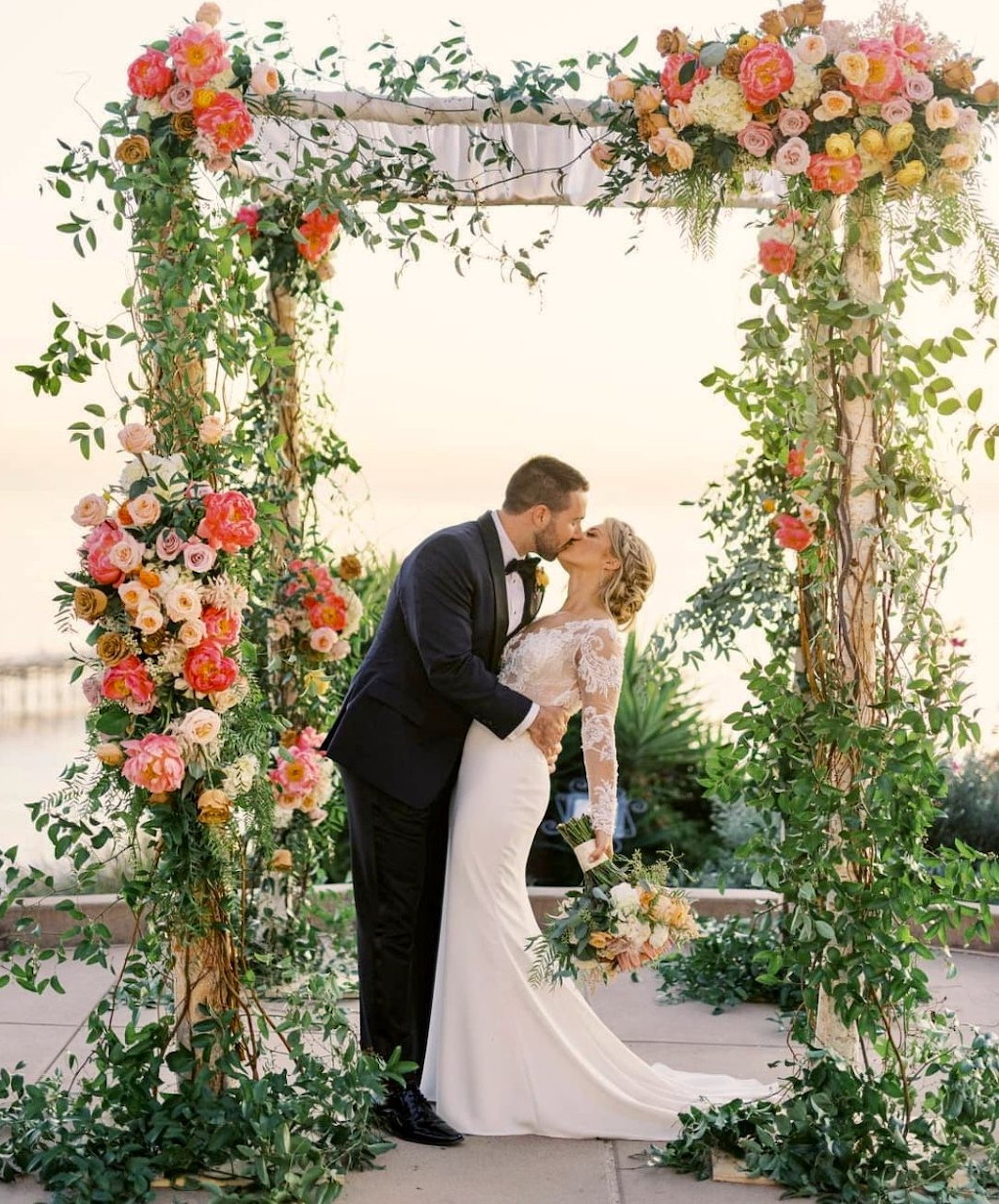 Bride and groom kissing under a flower-covered wedding arbor at sunset