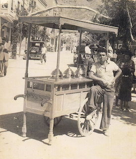 Vintage black and white photo of a man sitting next to a wooden ice cream cart on a street