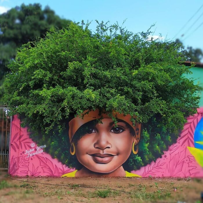 Street art mural of a face with lush green foliage as hair, pink wings, on turquoise wall