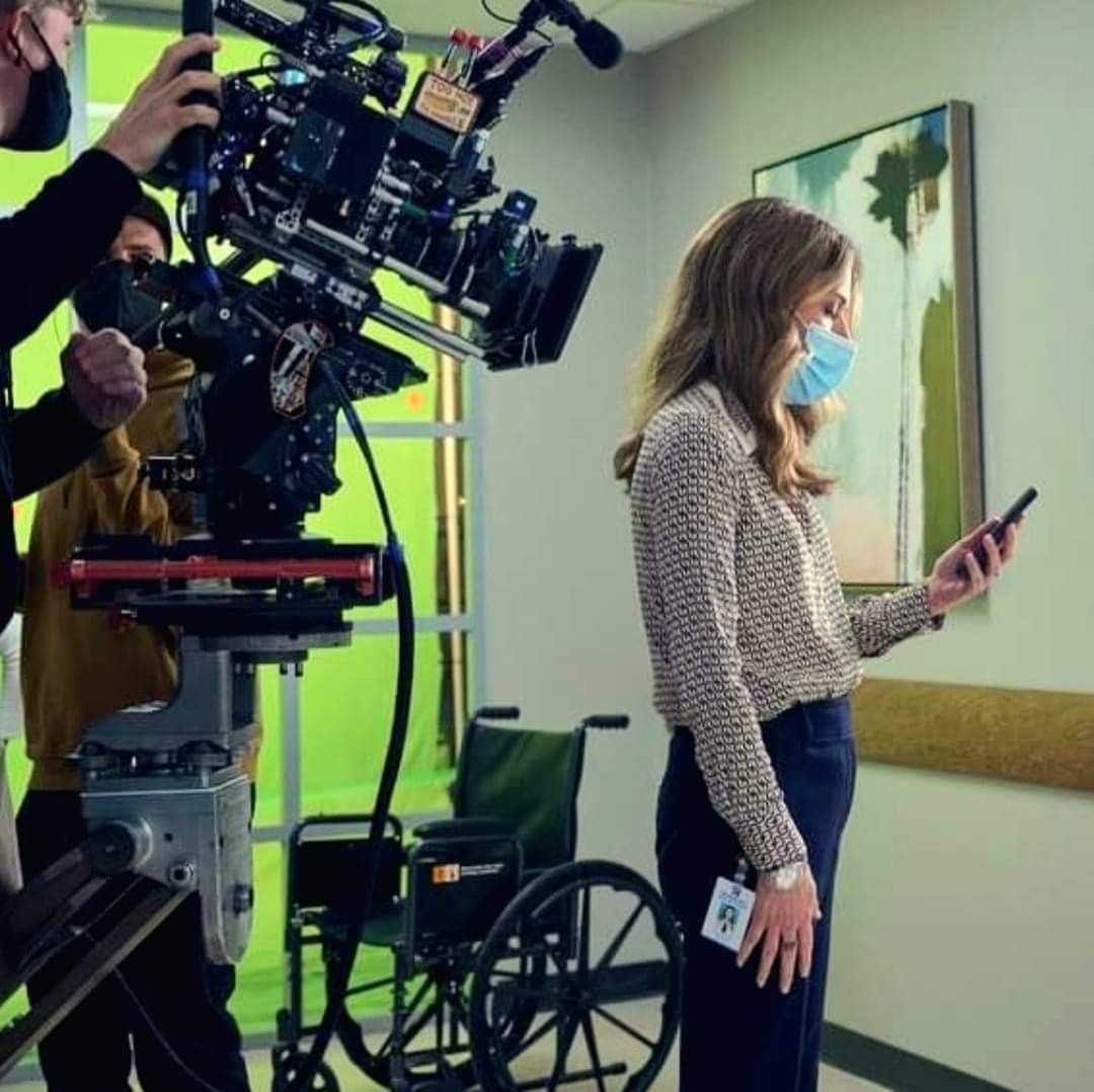Masked woman beside a camera rig and wheelchair in an indoor hallway