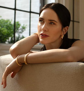 Dark-haired woman in black top leaning on cream sofa wearing gold earings and bracelets