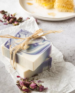 Handcrafted soap bar with purple swirls tied with raffia ribbon on white fabric beside dried flowers
