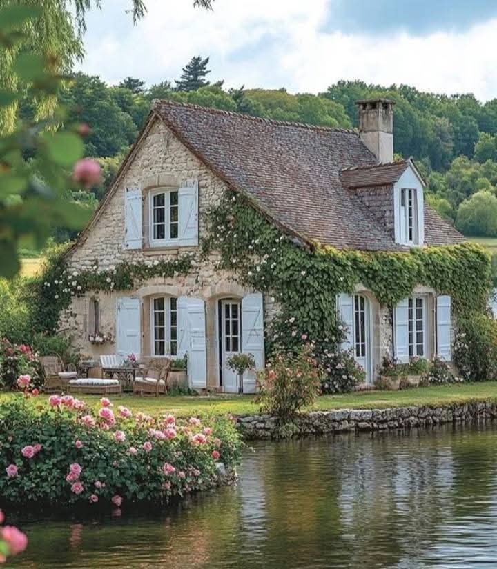 Stone cottage with blue shutters and climbing vines beside a calm pond surrounded by pink flowering bushes