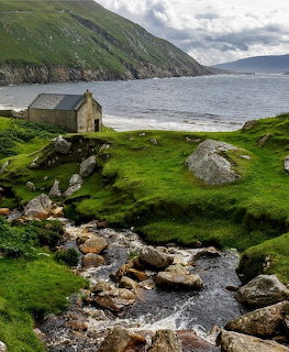 Stone cottage by a rocky stream on a green coastal hillside beside calm water