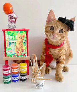 Orange tabby cat in beret and red scarf next to miniature art supplies and small cat painting
