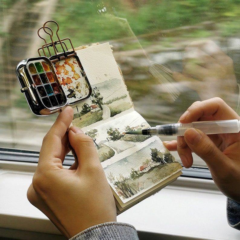 Hands painting a tiny landscape in a sketchbook with a small watercolor palette by a window