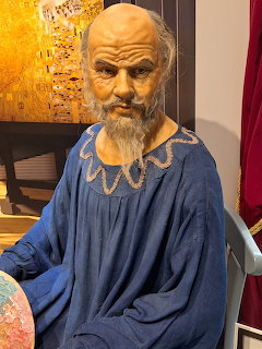 Wax figure of bearded man in blue robe with decorative neckline seated near golden artwork
