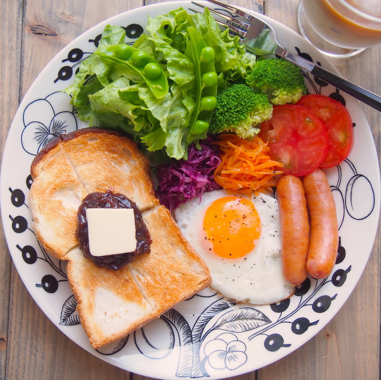 Overhead view of a breakfast plate with toast, egg, sausages, and assorted fresh vegetables on a decorative plate
