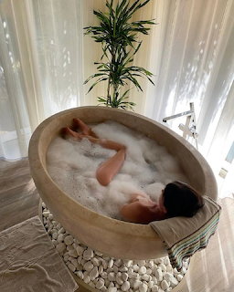 Woman relaxing in an oval stone bathtub filled with bubble bath, surrounded by white pebbles and curtains