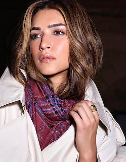Woman with brown hair in white jacket and purple plaid scarf posing against dark background
