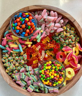 Wooden bowl filled with assorted colorful candies including gummies, chocolates, sour worms, and sprinkles