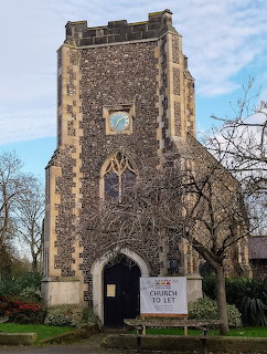 Stone church tower with flint facade, Gothic arches, and bare trees under blue sky