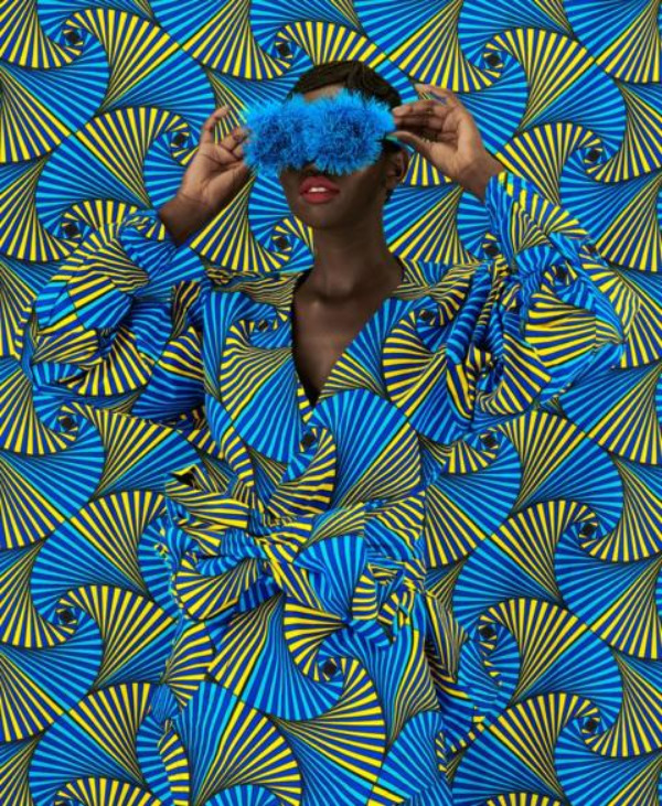 Person in blue and yellow African print outfit blending into matching patterned background, holding blue fur eye mask