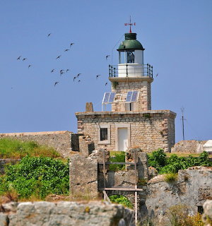 Stone lighthouse on a rocky hillside with birds flying in a clear blue sky