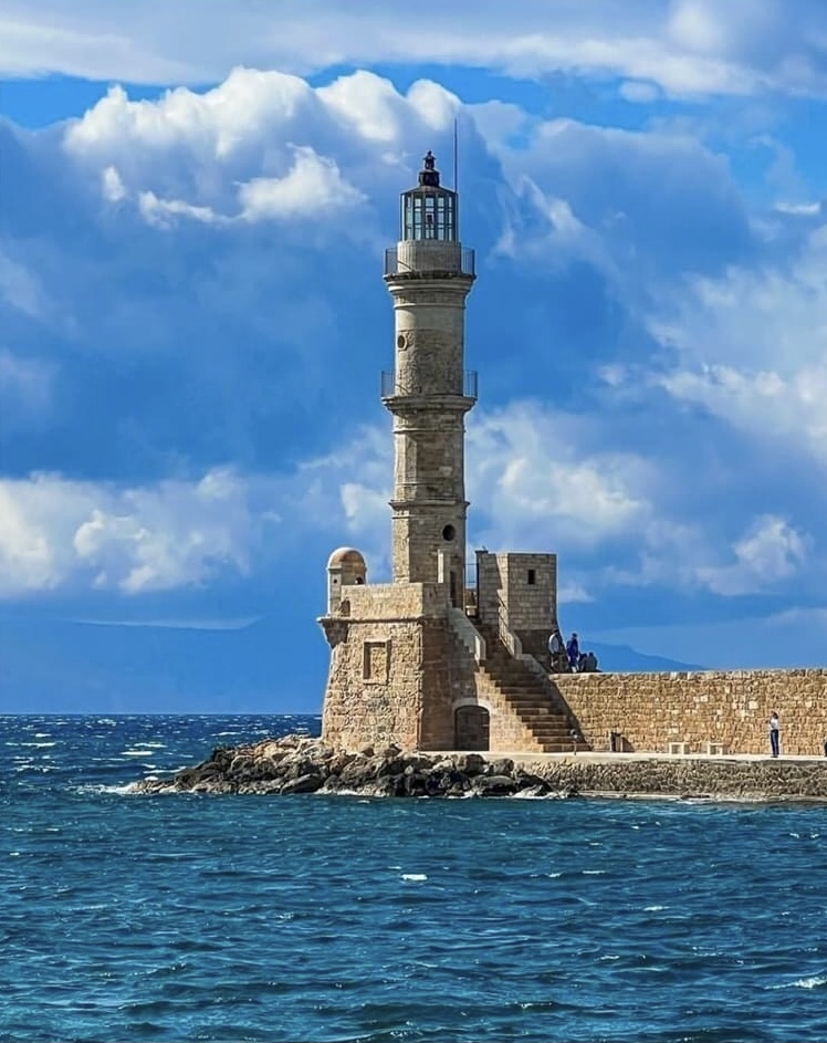 Tall stone lighthouse tower on rocky pier extending into blue sea under cloudy sky