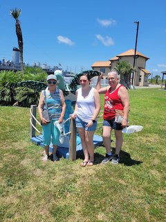 Three women posing on grass with palm trees and blue sky behind them