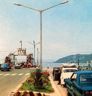 Vintage cars parked by a ferry dock on a waterfront road with hills in the distance