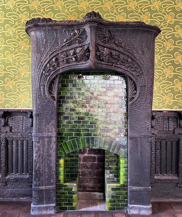 Ornate Art Nouveau stone fireplace with carved details and green glazed brick interior against yellow wallpaper