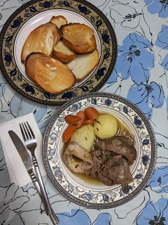 Two blue-rimmed plates on a floral tablecloth, one with boiled meat and vegetables in broth, one with bread rolls.