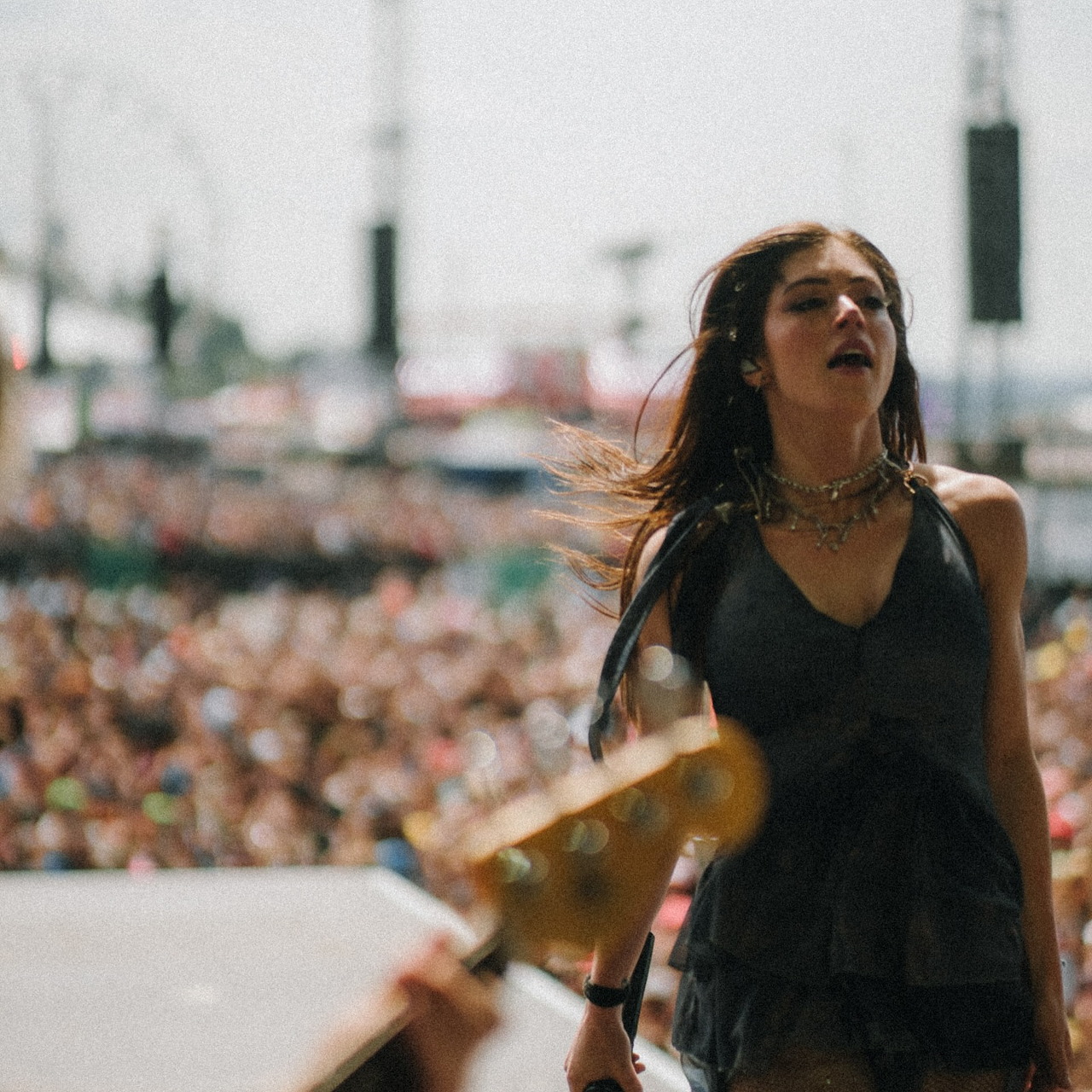 Female singer on an outdoor stage with a crowd behind her and a blurred guitar in the foreground