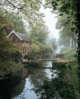 Brick house beside a quiet canal with trees, reflections, and light mist