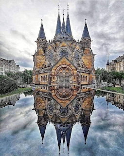 Ornate castle with tall spires reflected symmetrically in water under cloudy sky