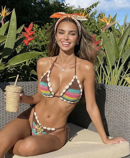 Smiling brunette woman in colorful crochet bikini sitting outdoors with tropical plants behind her