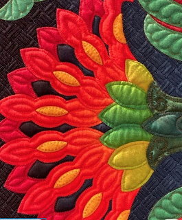 Close-up of a colorful quilted flower with red petals, yellow accents, and green leaves on dark fabric