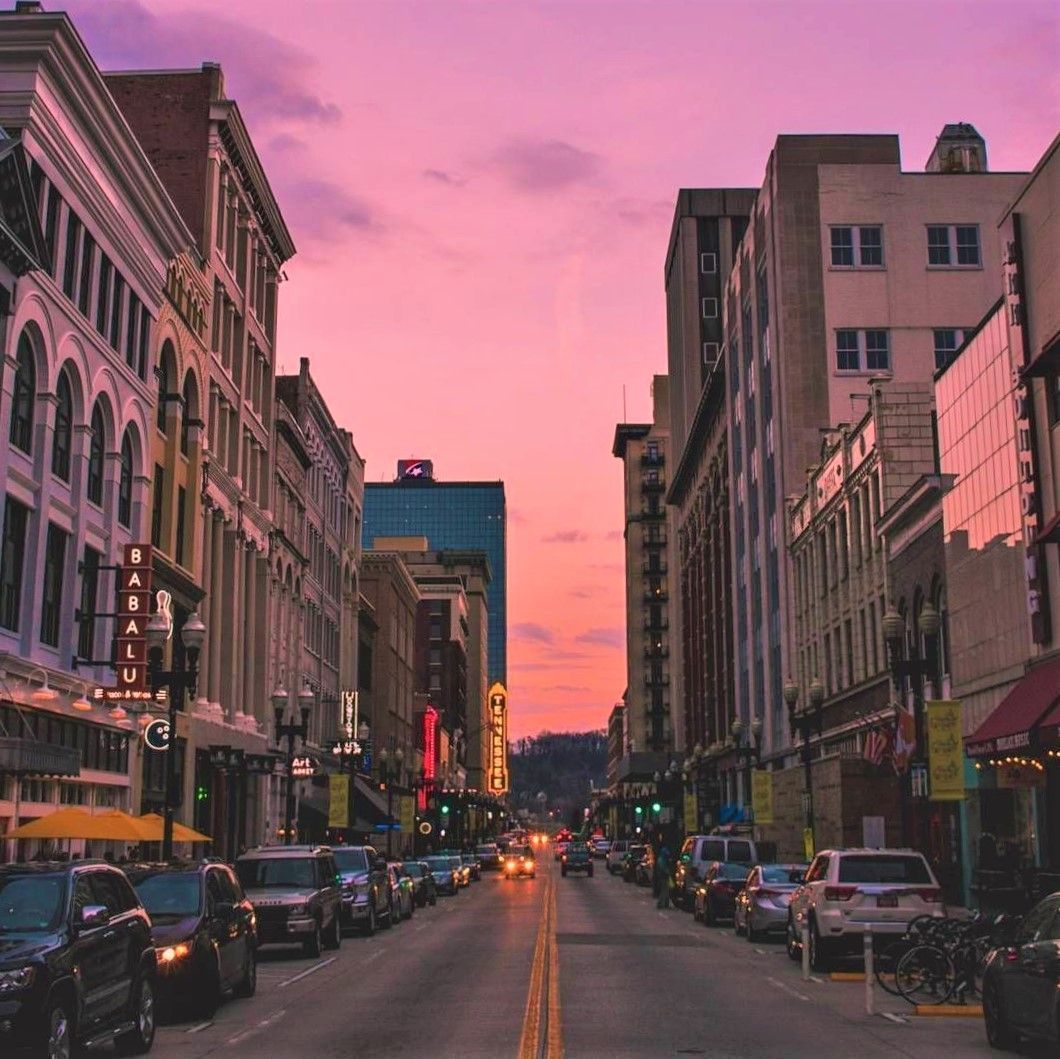 Downtown street with cars and tall buildings beneath a pink sunset sky