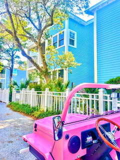 Pink open-top jeep parked in front of a bright blue beach house with white fence and tropical trees