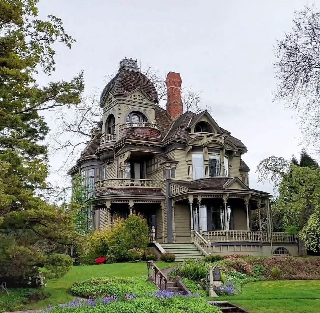 Gray Victorian mansion with tower and wraparound porch surrounded by trees and lawn