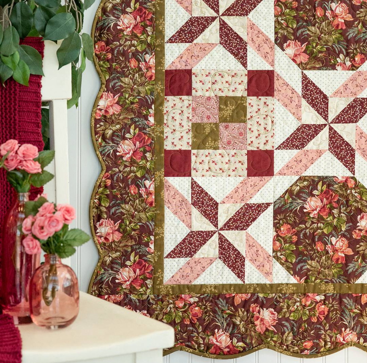 Patchwork quilt with burgundy and pink star pattern blocks and floral fabric borders draped beside vase of pink roses