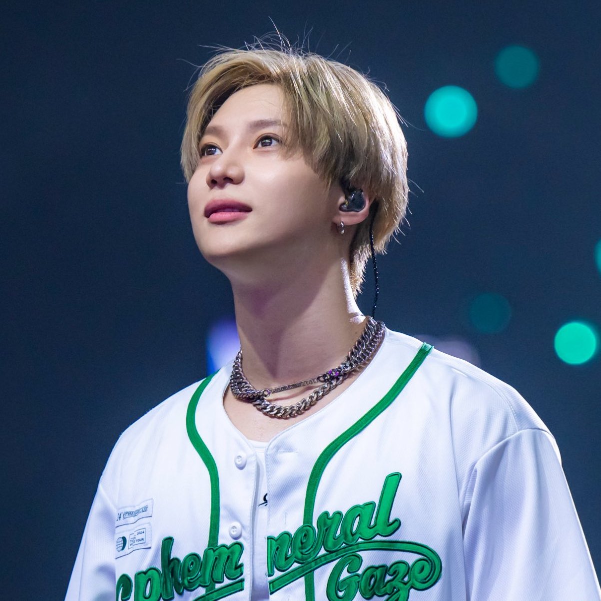 Close-up of Taemin wearing a white jersey and necklaces on stage, looking upward
