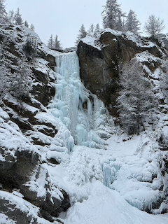 Frozen waterfall on rocky cliff face surrounded by snow-covered rocks and evergreen trees in winter