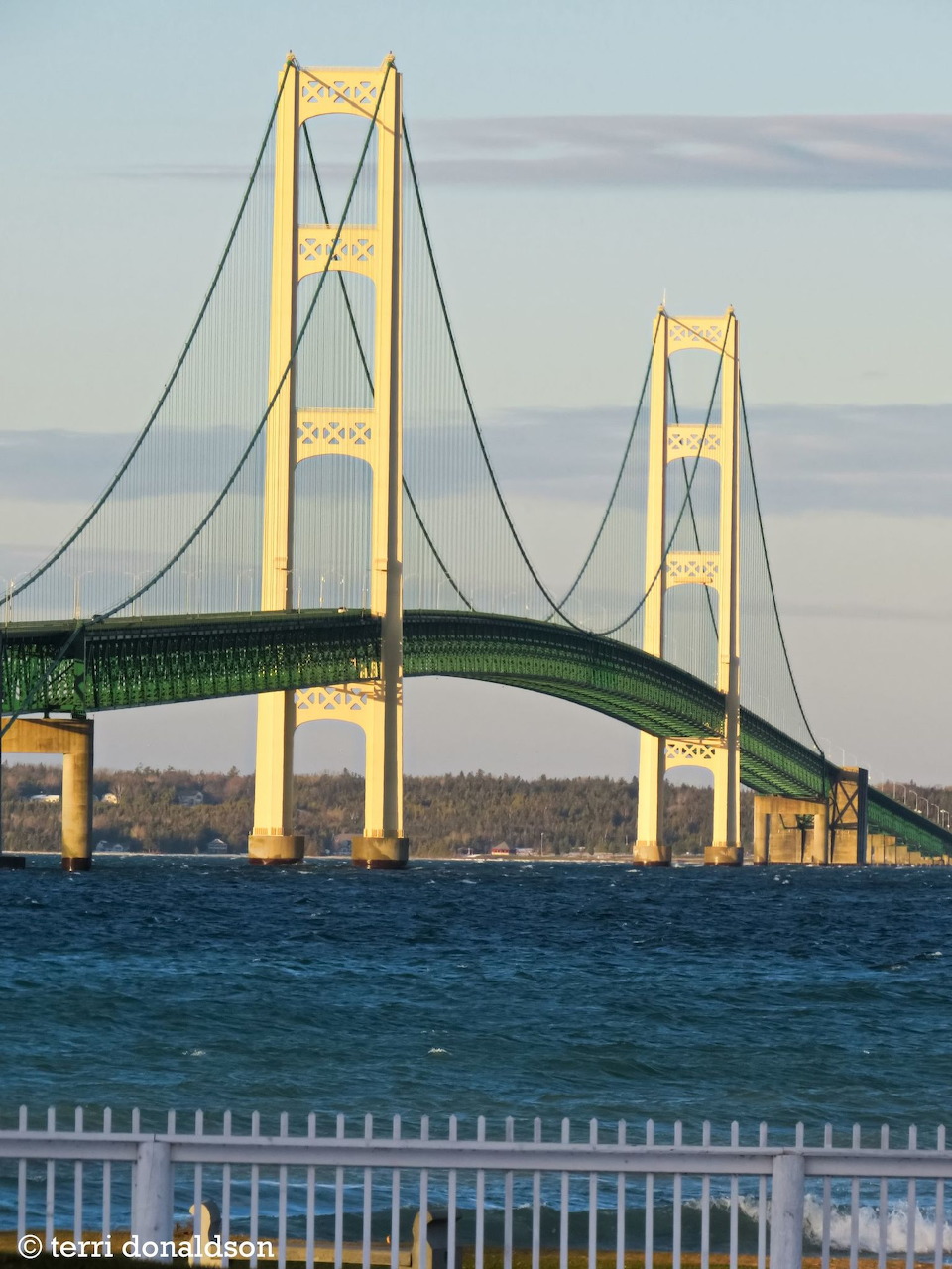Mackinac Bridge over blue water with a white fence in the foreground
