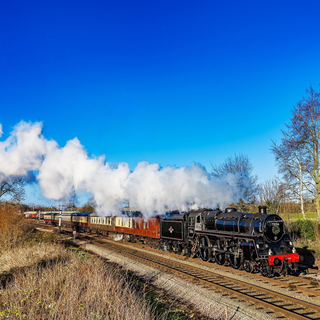 Black steam locomotive pulling freight cars on tracks with white steam against blue sky