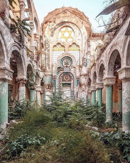 Ruined interior of abandoned synagogue with arches, ornate window, and overgrown plants