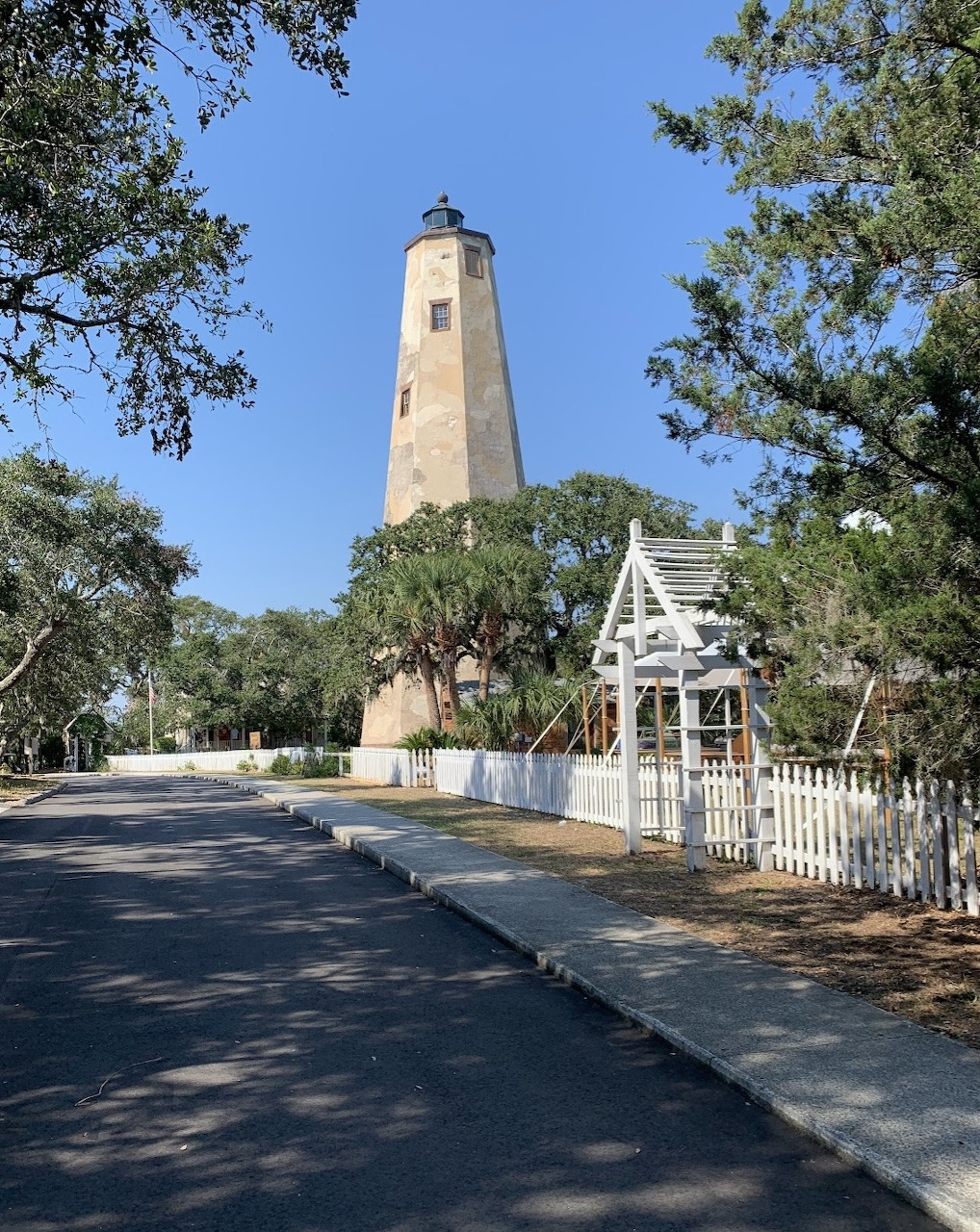 Tall stone lighthouse surrounded by oak trees with white picket fence and arbor in foreground