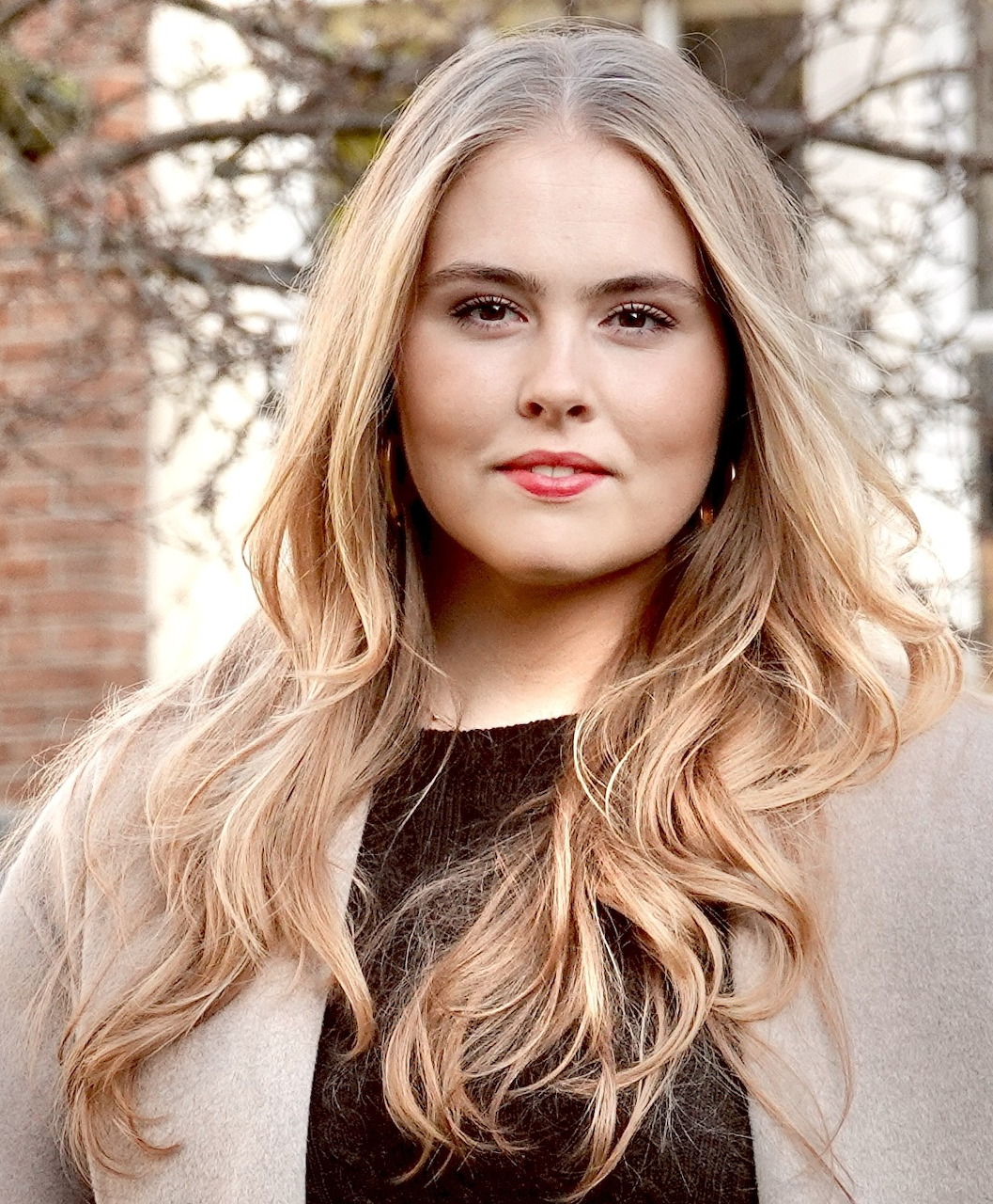 Young blonde woman with long wavy hair in a dark top and beige coat posing outdoors