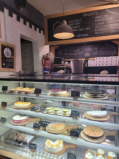 Glass display case in a café showing multiple whole cakes and desserts with a chalkboard menu behind the counter.