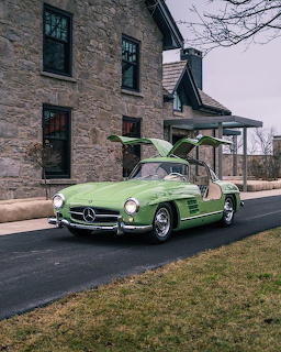Light green Mercedes-Benz 300 SL Gullwing with doors open on a driveway beside a stone building.