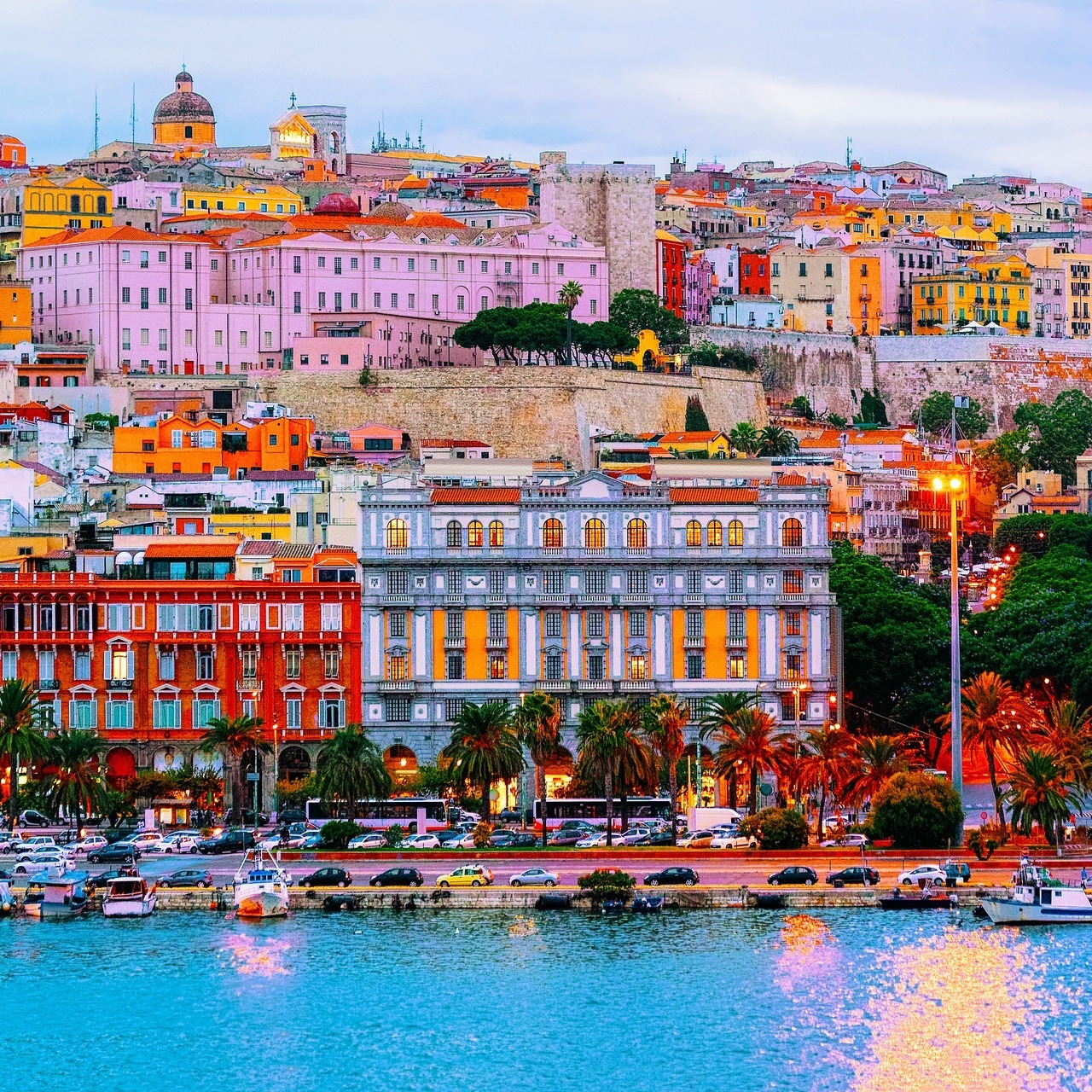 Historic city buildings along a waterfront with palm trees, boats, and blue water at dusk