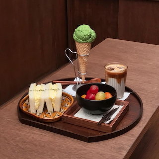 Tray with layered iced coffee, green ice cream cone, fruit bowl, and small sandwiches on a wooden table