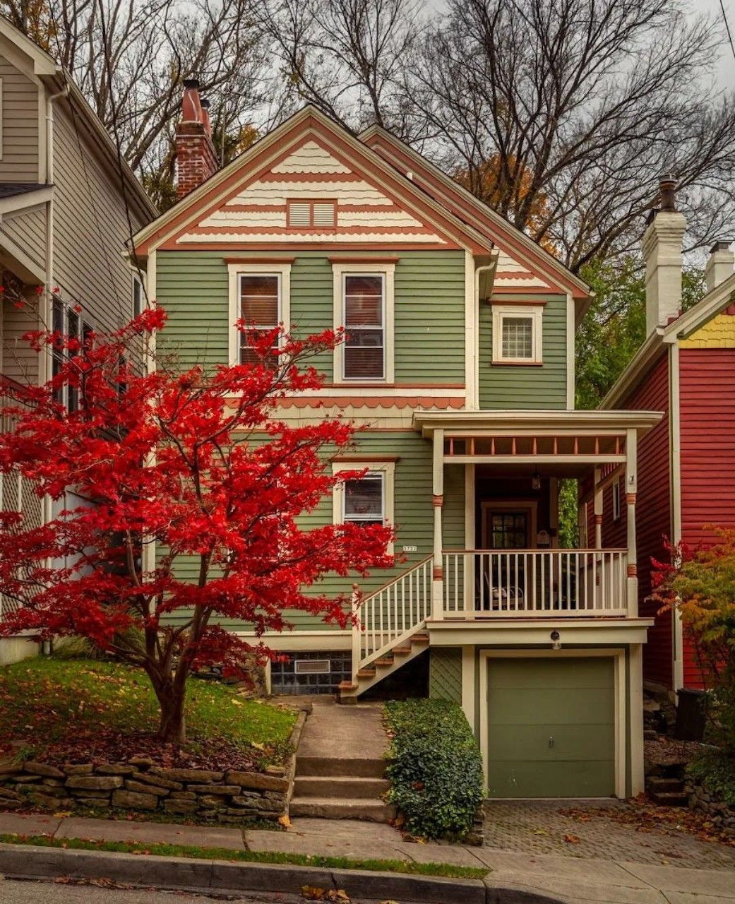 Green two-story house with red maple tree in front yard on residential street