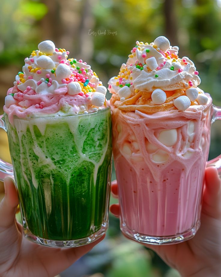 Two hands holding colorful milkshake glasses topped with whipped cream, sprinkles, and marshmallows
