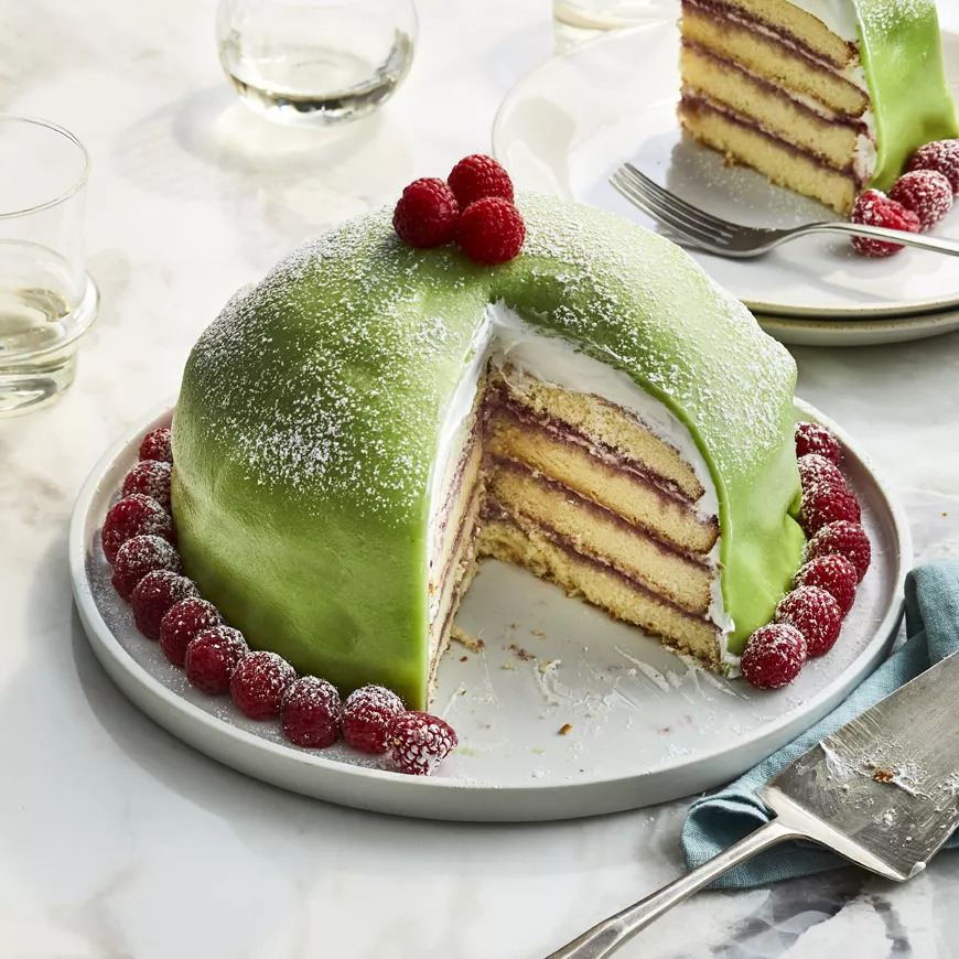 Green dome-shaped princess cake with raspberry garnish, one slice cut to show cream and jam layers inside
