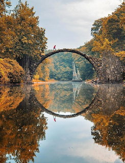 Stone arch bridge reflected in calm water forming a perfect circle, framed by autumn trees