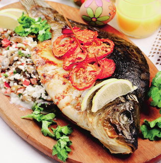 Whole baked carp on a wooden board with rice stuffing, tomato slices, lemon wedges, and herbs