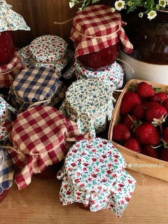 Several jam jars with fabric lids in gingham and floral prints next to a basket of fresh strawberries on a wooden surface.